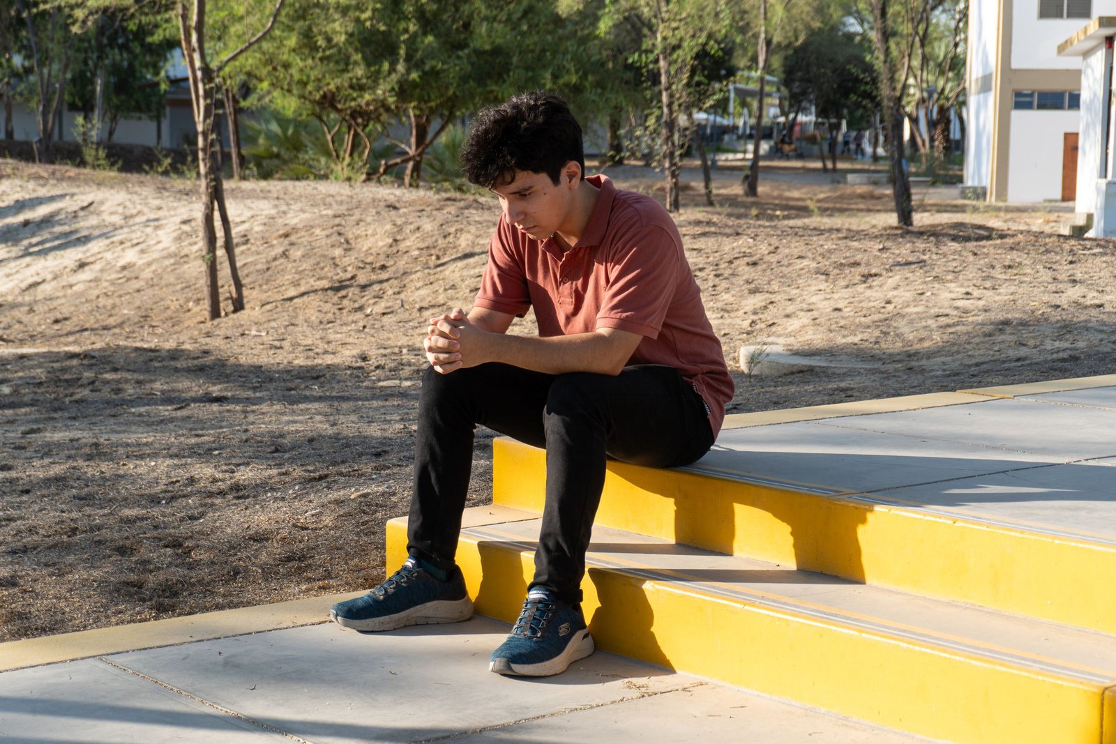 A young man sits on yellow steps in a park in Piura, Peru, lost in thought during a sunny day.